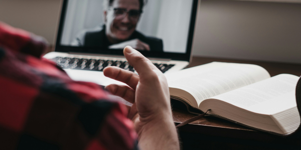 A person gestures to another person on a laptop screen near an open book.